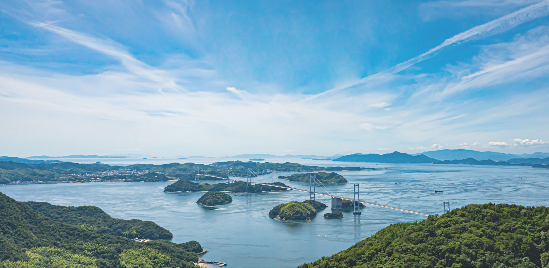 Photo: The beautiful Shimanami Sea Route with blue skies. Several small, lush green islands dot the center, and a large bridge can be seen connecting them.