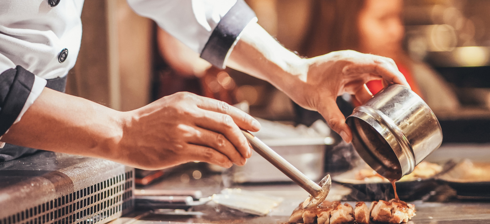 Photo: Chef’s hand at work cooking. The chef uses a stainless steel container and spoon to pour the sauce over the food.