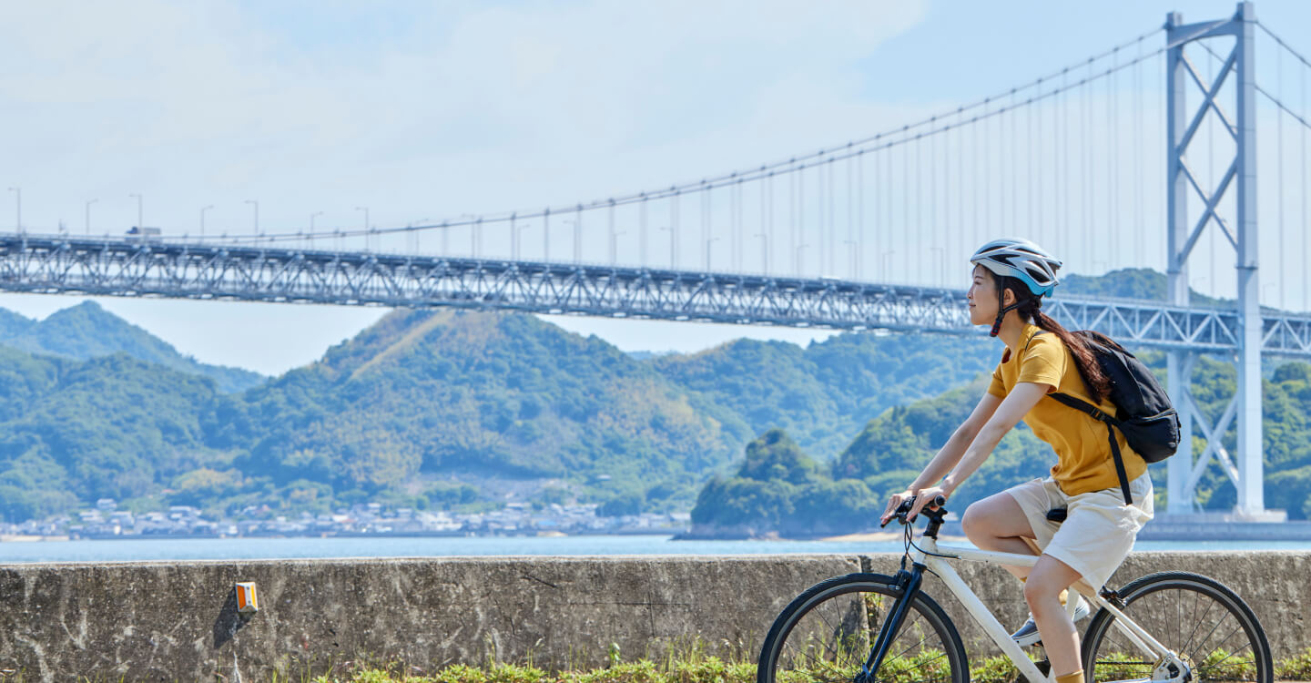 写真：自転車に乗る女性が、晴れた日、しまなみ海道の海沿いを走り、背景には大きな橋と山々が広がっている