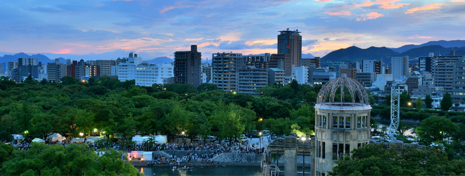 写真：背景には山々があり、マンションやビルが沢山建っている夕暮れの広島市内と、たくさんの観光客が集まっている原爆ドーム