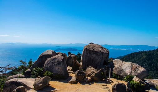 写真：大きな岩が点在する頂上からは、遠くに広がる海と山々が一望でき、澄んだ青空が広がる弥山展望台