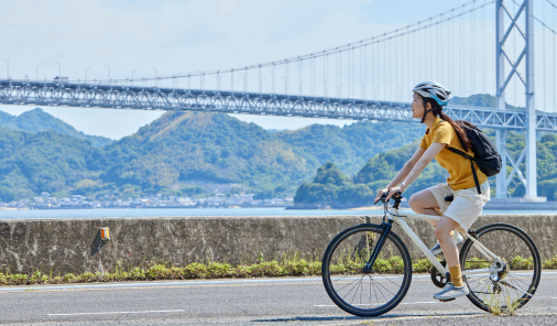 写真：自転車に乗る女性が、晴れた日、しまなみ海道の海沿いを走り、背景には大きな橋と山々が広がっている